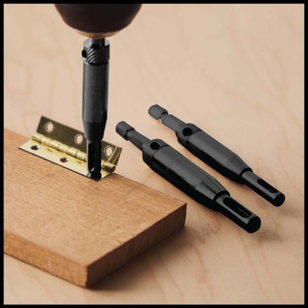 Three black metal screwdriver bits are shown on a light wood surface, with one bit being used to drive a brass hinge into a wooden block. A drill chuck is also visible at the top.