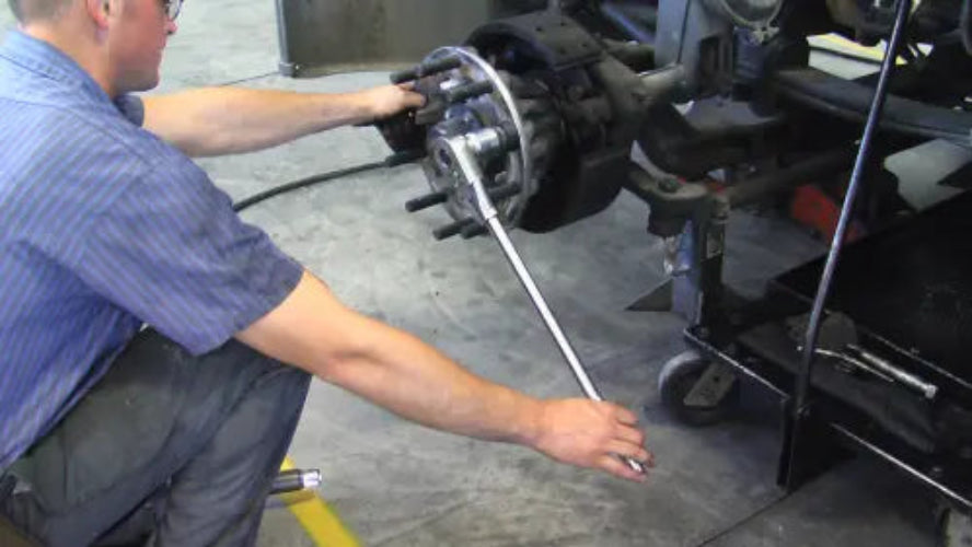 A mechanic wearing a blue striped shirt uses a long wrench to loosen a large wheel hub on a vehicle axle in an auto repair shop.