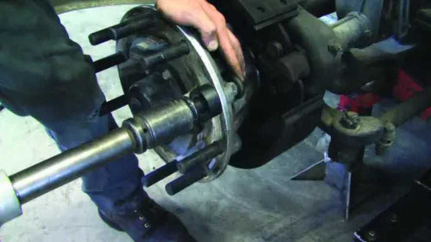 A close-up of a person's hand and legs working on a vehicle wheel hub assembly mounted on a metal spindle in a workshop setting.