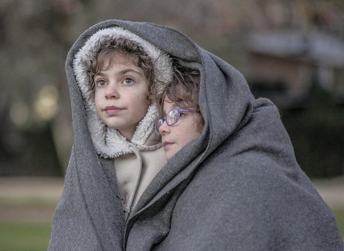 Two children wrapped in a blanket outdoors on a cold day.