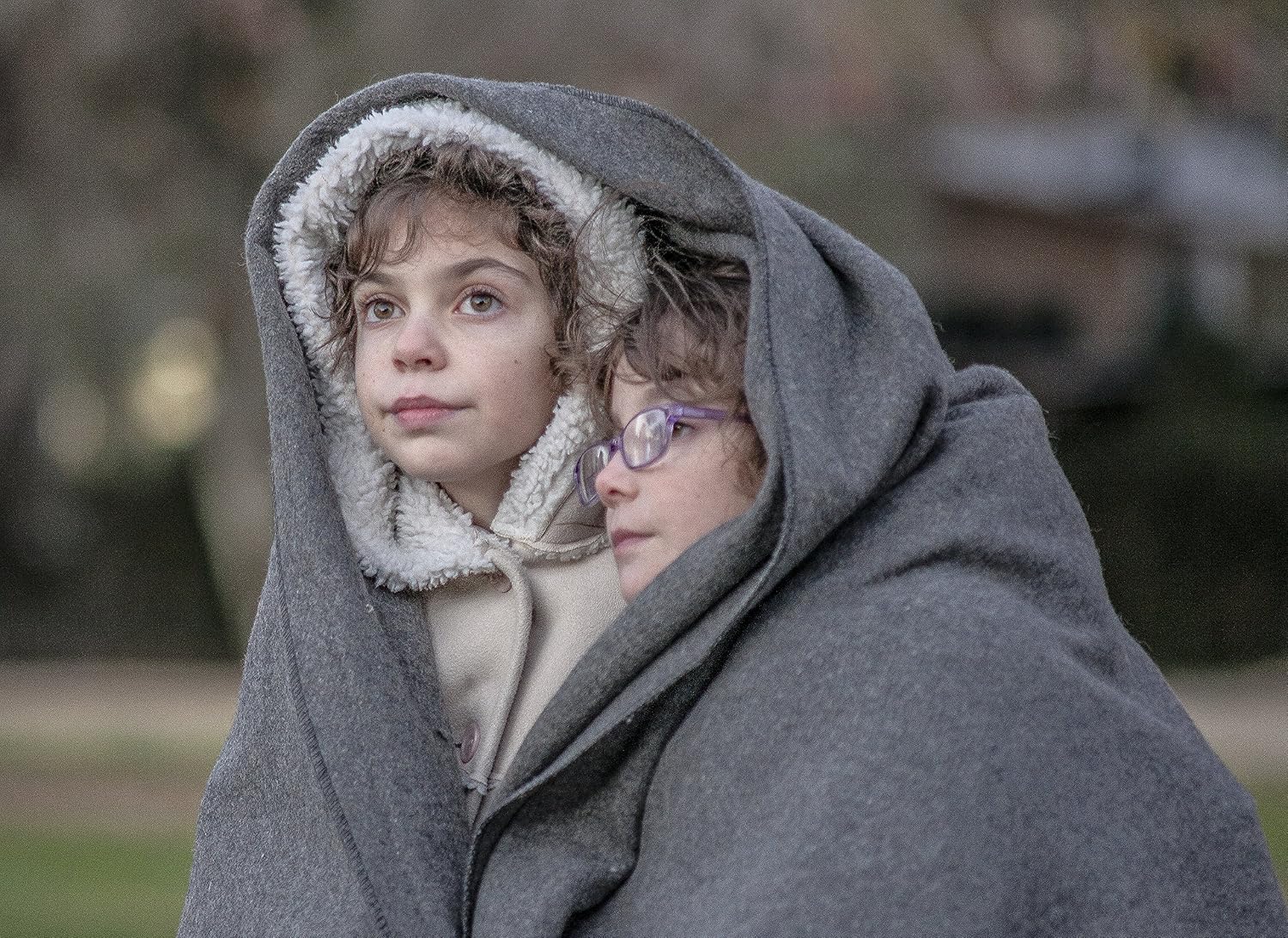 Two children wrapped in a blanket outdoors on a cold day.