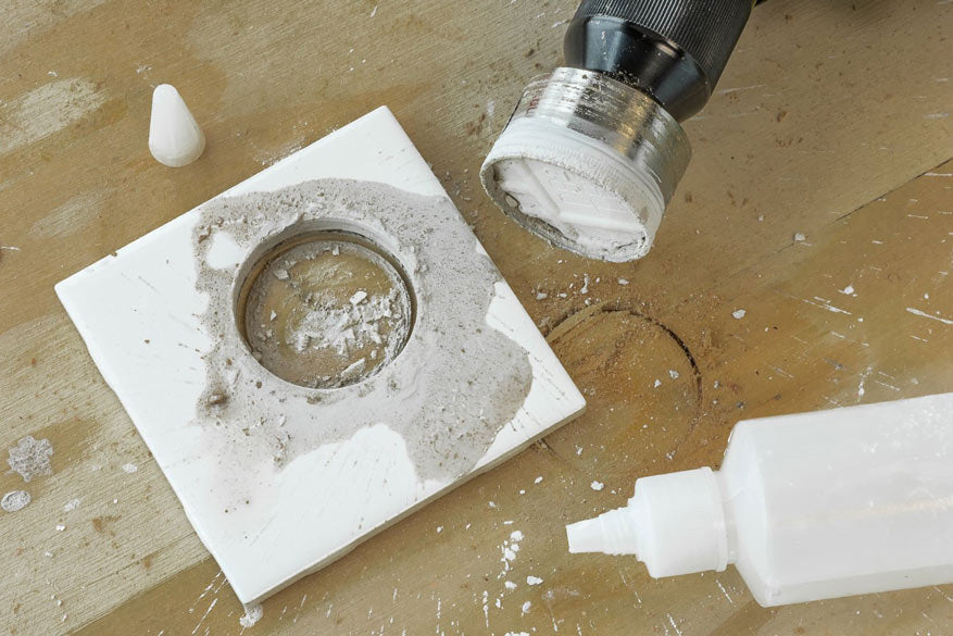 A close-up of a circular tile or stone insert with a rough textured center, shown on a workbench next to a bottle of adhesive, a small white cone-shaped piece, and a drill attachment.
