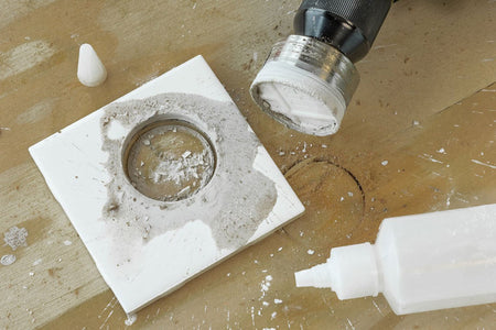 A close-up of a circular tile or stone insert with a rough textured center, shown on a workbench next to a bottle of adhesive, a small white cone-shaped piece, and a drill attachment.