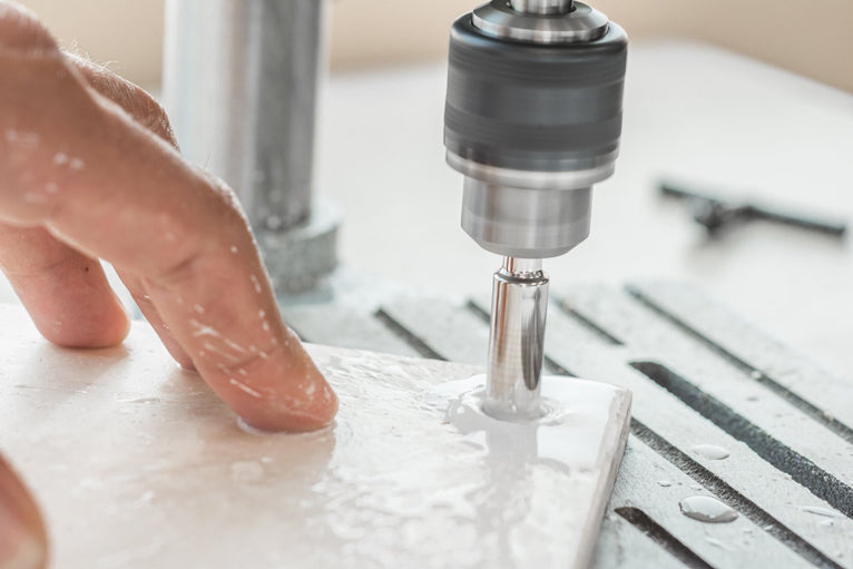 A close-up action shot of a person's hand guiding a white ceramic tile while a drill press with a metal burr bit drills a hole into it. The scene is set on a metal workbench with a wet, dusty workspace background.