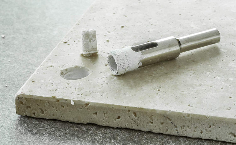 A metal hole saw drill bit with a white protective coating and a small white cylindrical accessory, shown resting on a light gray concrete tile over a darker gray surface.