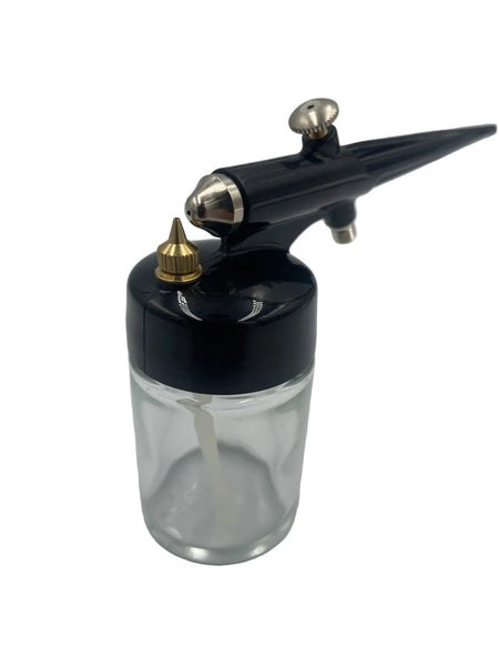 A black airbrush paint gun with silver metal accents and a gold nozzle, attached to a clear glass ink bottle, shown on a light gray background.